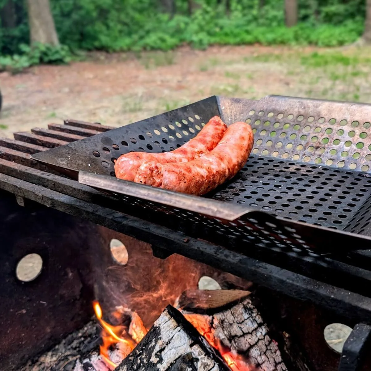 Sausages cooking in a perforated stainless steel grill basket set over an open campfire