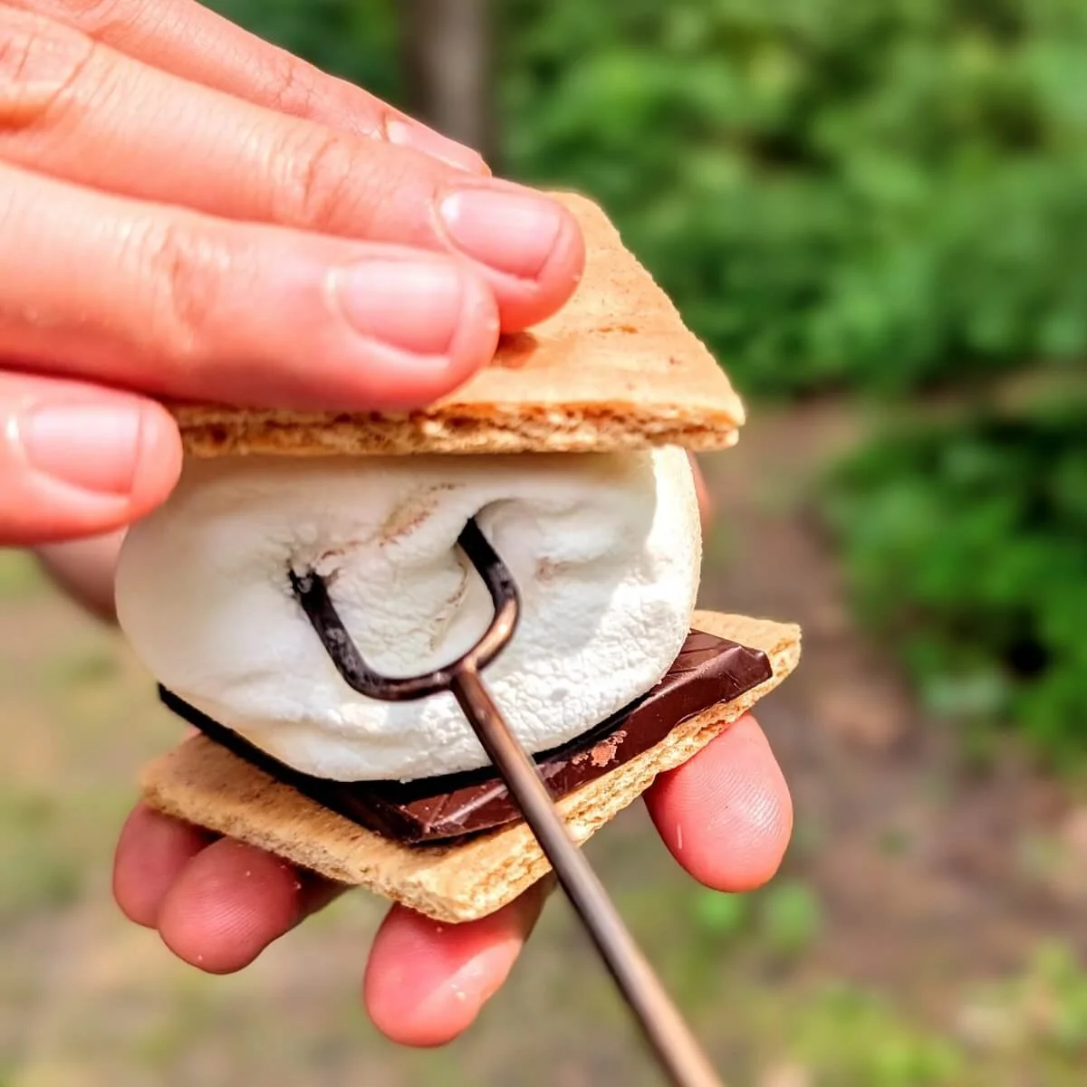Roasted marshmallow on a telescoping camp fork being pressed into a s’more with graham crackers and chocolate at a campsite