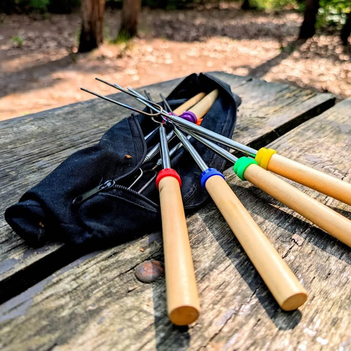 Telescoping campfire roasting forks with wooden handles and colored rings resting on a camp table beside a black zippered storage pouch