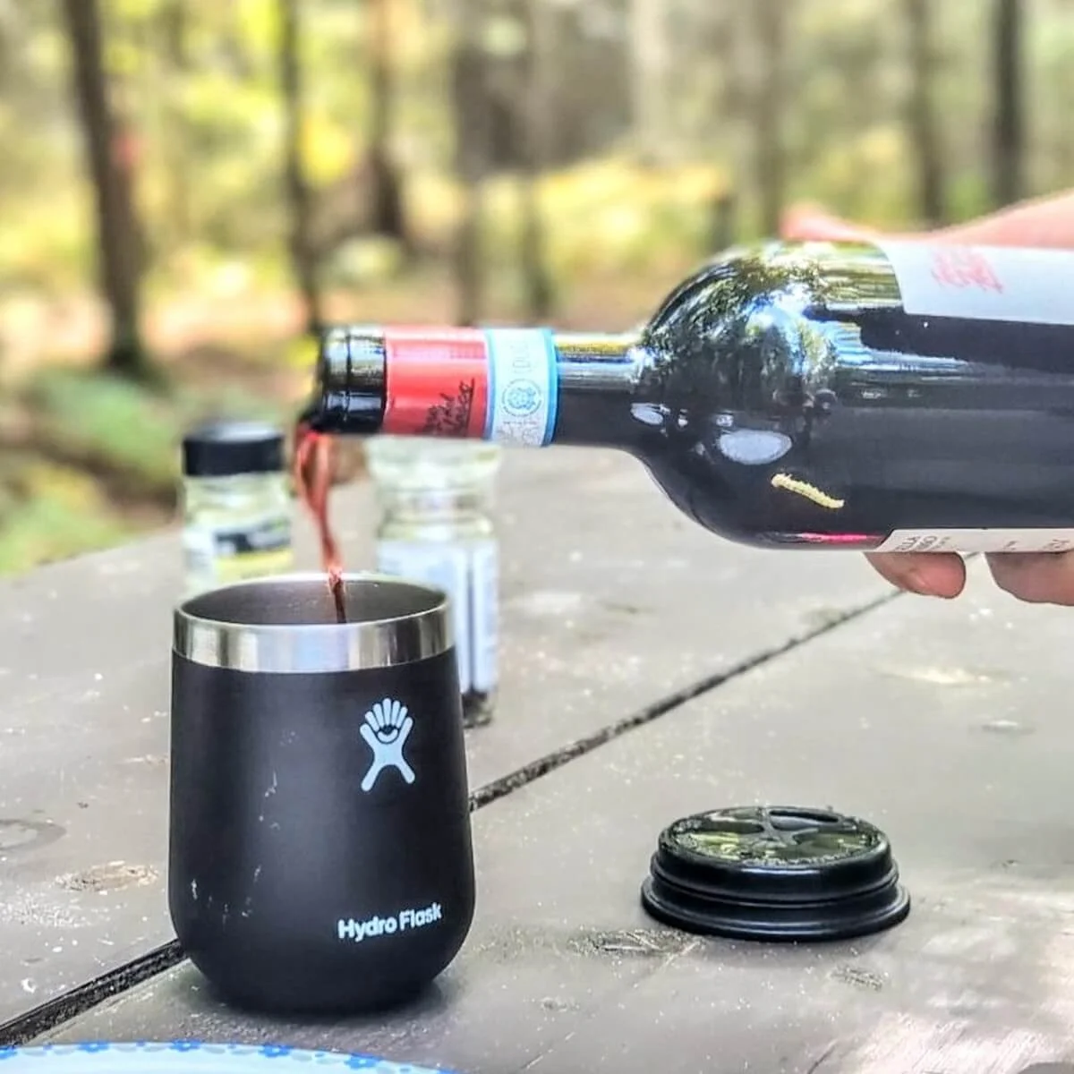 Wine being poured into insulated travel wine tumbler at campfire campsite picnic table
