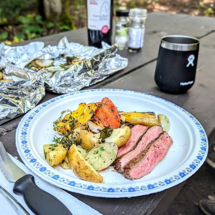 Campfire meal with sliced steak roasted vegetables and insulated travel wine tumbler on picnic table