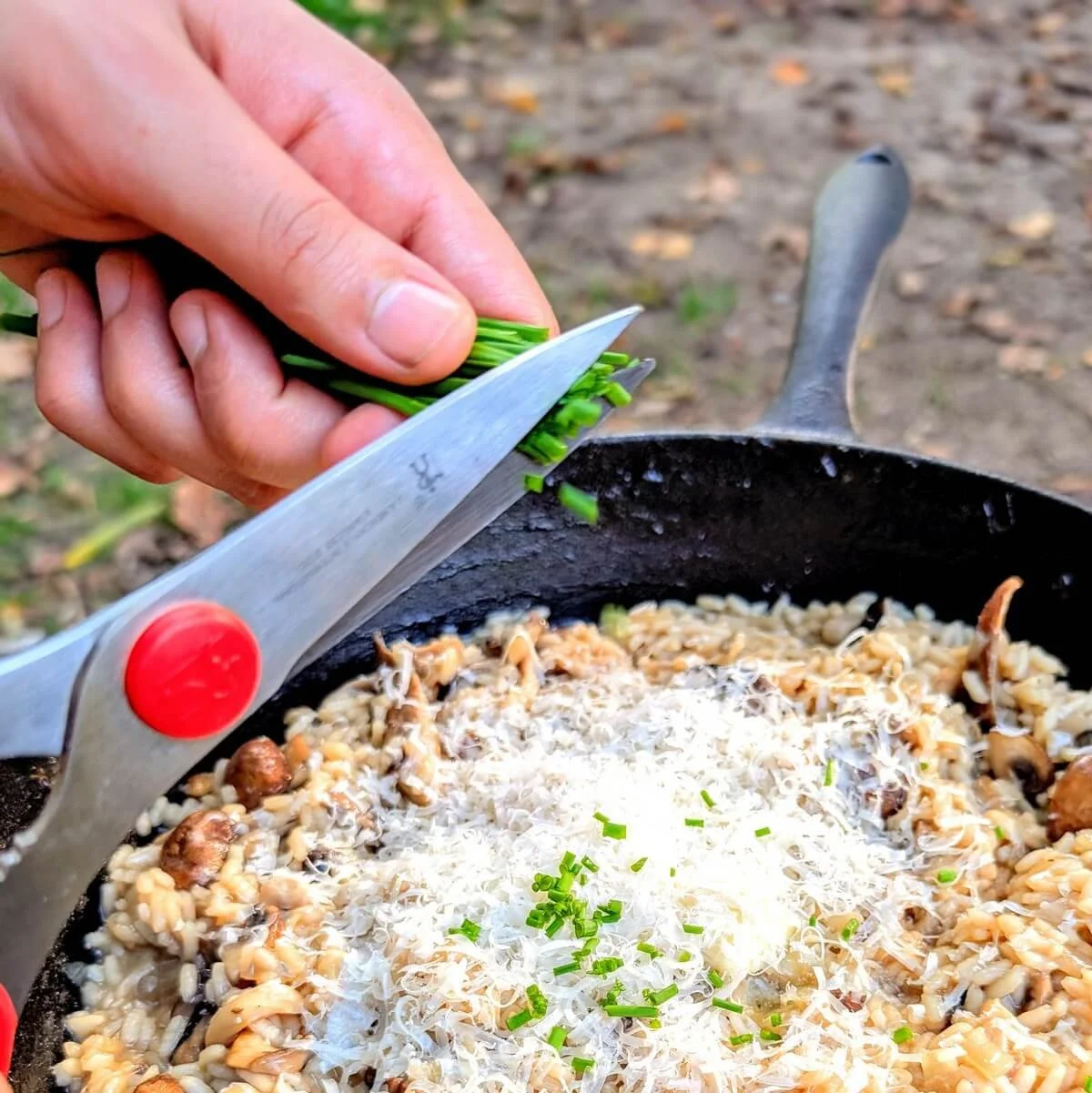 Kitchen scissors cutting fresh chives over campfire risotto in cast iron pan