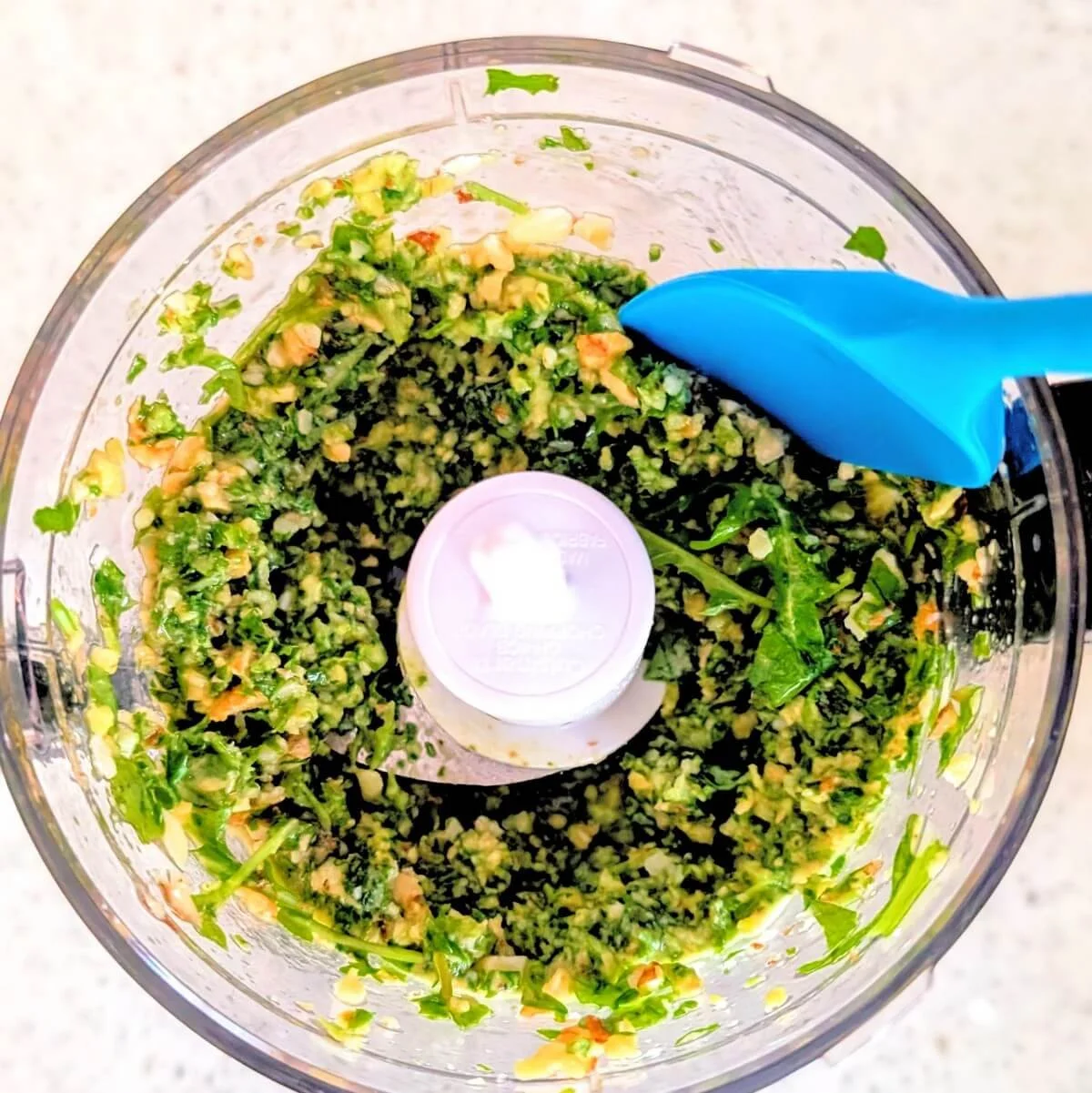 Spatula scraping down the sides of a food processor while making pesto