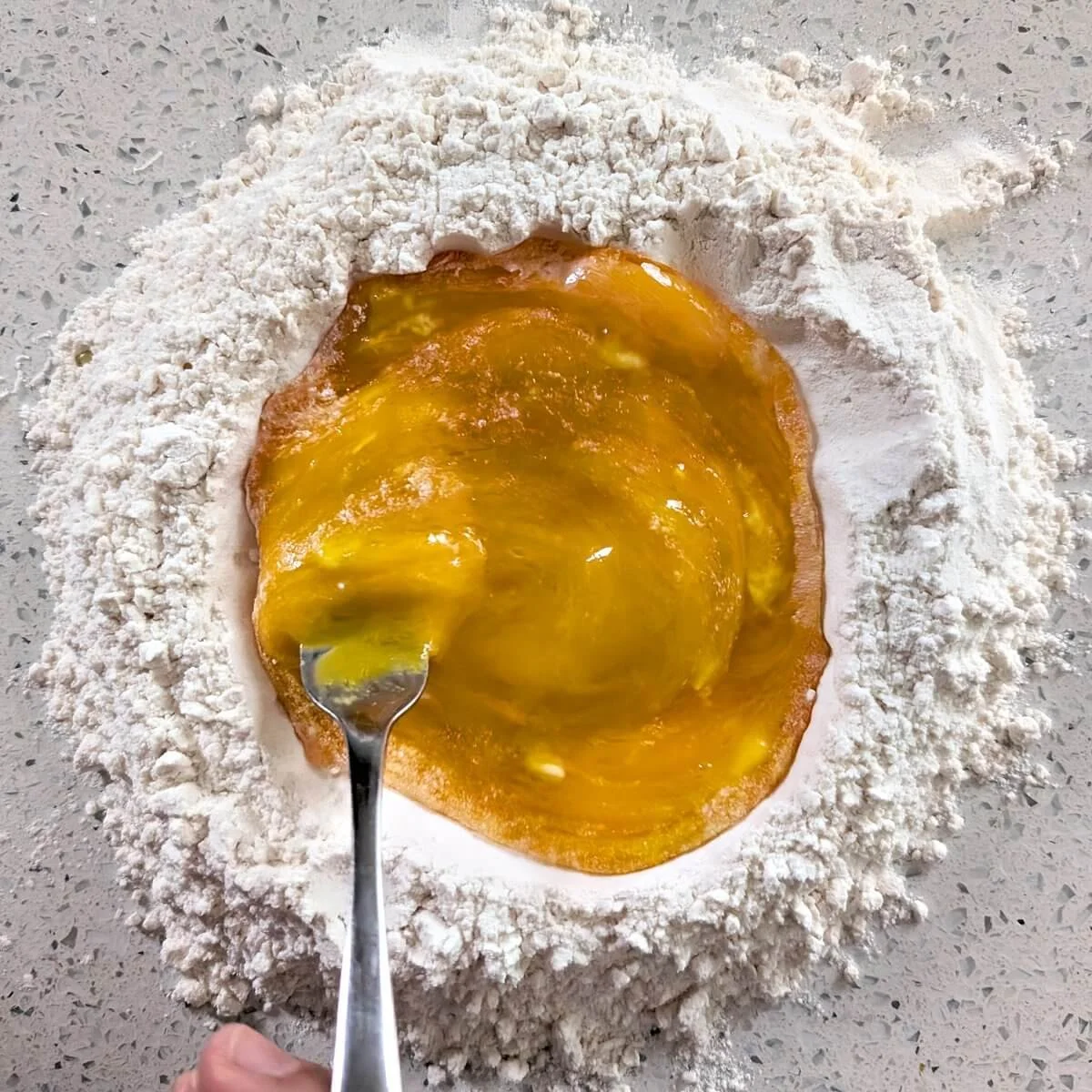 Eggs and yolks being whisked with a fork inside a flour well on a countertop to make fresh pasta