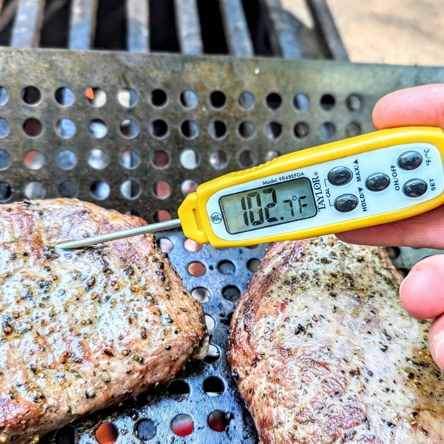 Digital meat thermometer showing 102.7°F in flank steak over open fire