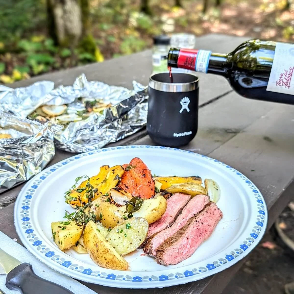 Grilled camping steak on a paper plate with vegetables and wine being poured in the background