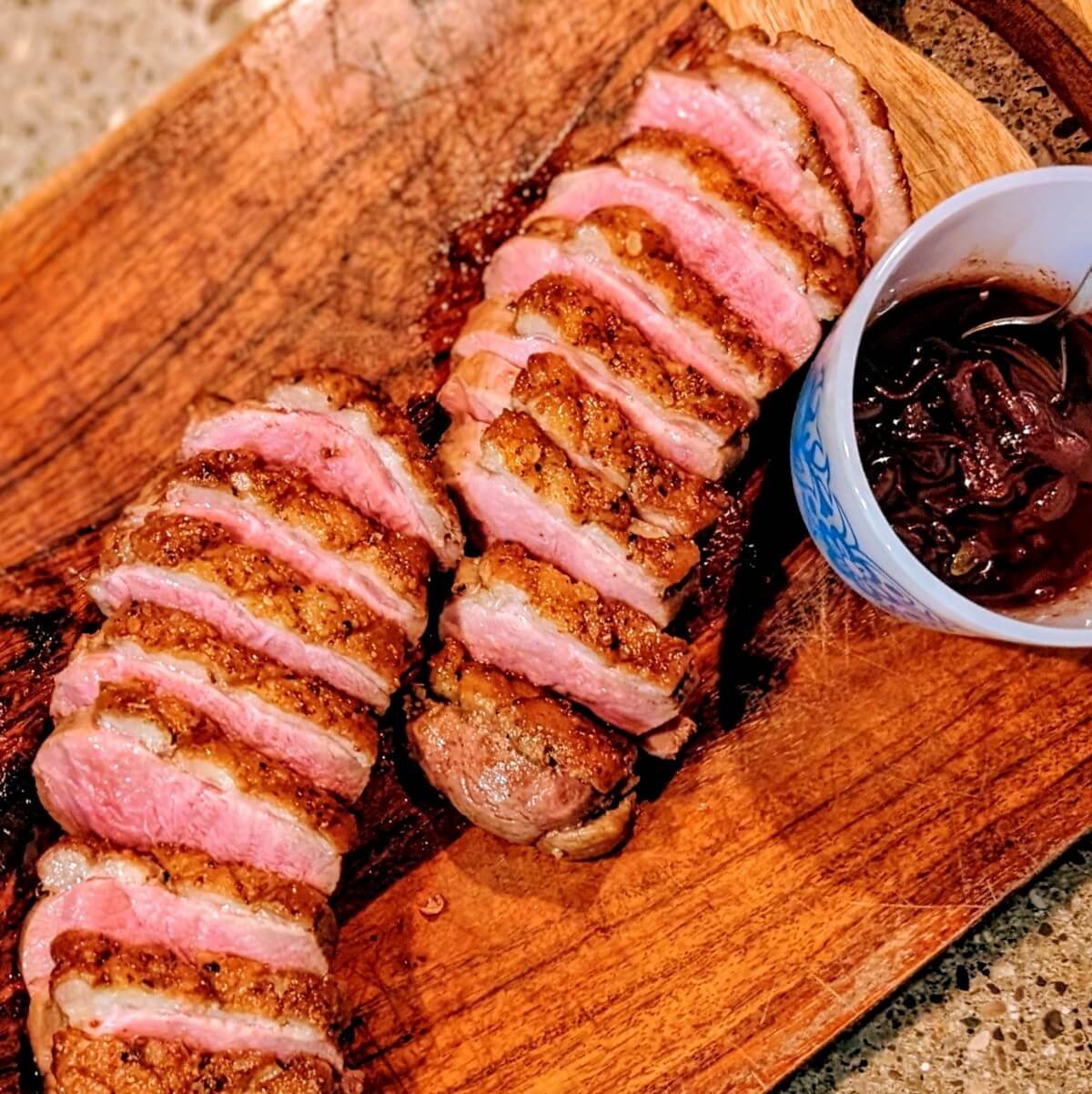 Sliced crispy duck breast on a wooden board with red wine pan sauce in a small bowl