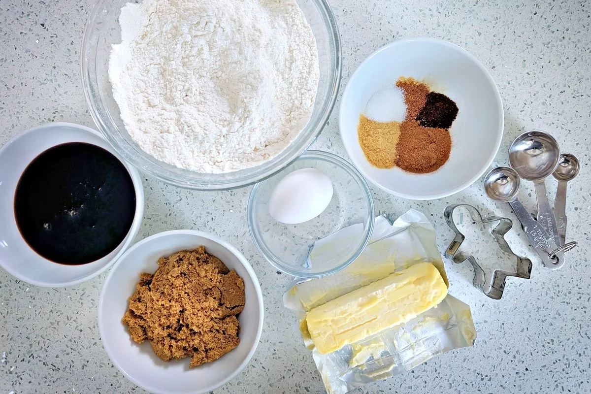 Measured gingerbread cookie ingredients with flour, spices, molasses, brown sugar, egg, butter, cookie cutters, and measuring spoons