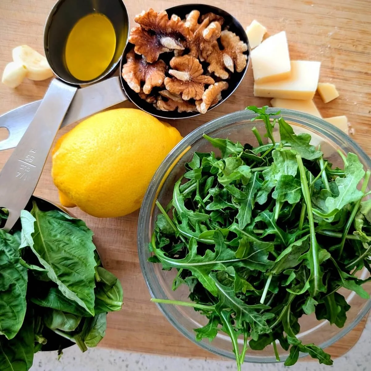 Baby arugula, fresh basil, lemon, walnuts, Parmesan, garlic, and olive oil arranged on wooden cutting board for arugula basil pesto recipe