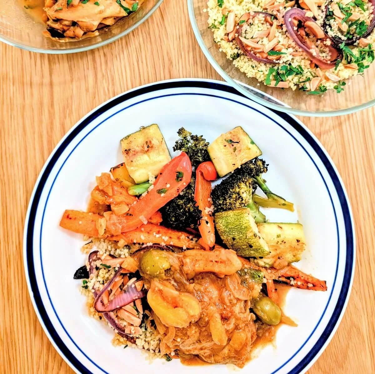 Plate filled with kettle couscous, chicken tagine, and roasted vegetables with serving bowls of couscous and tagine on a wooden table