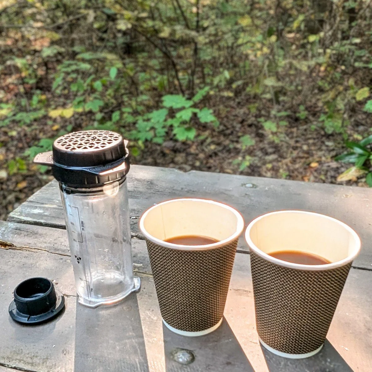 Delter Coffee Press on a campsite picnic table beside two cups of freshly brewed coffee