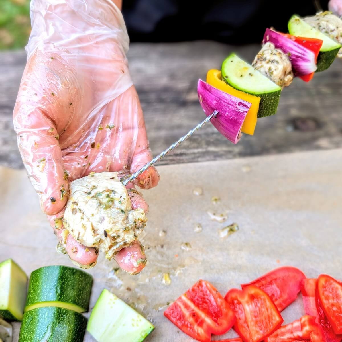 Gloved hand threading marinated chicken, zucchini, red onion, and bell pepper onto metal skewers for campfire grilling