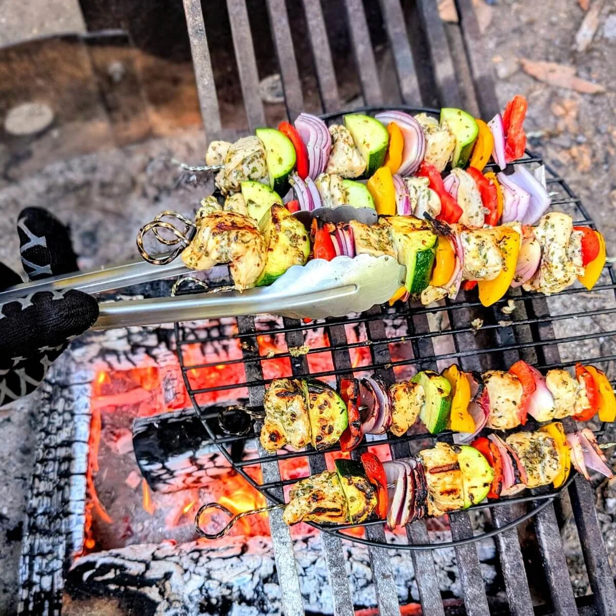 Gloved hand using tongs to rotate chicken and veggie skewers on the grill over a campfire