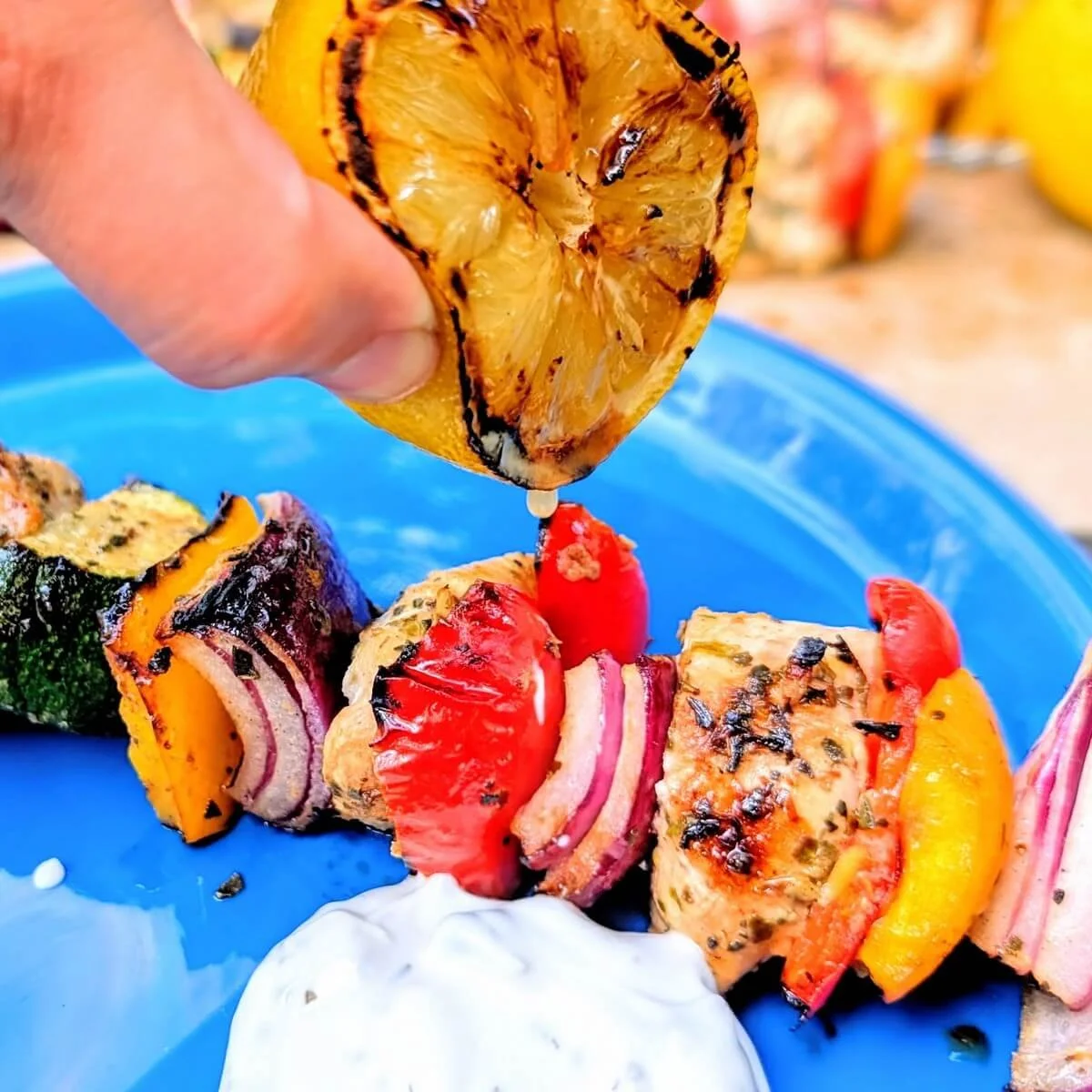 Grilled lemon being squeezed over Greek chicken skewers with tzatziki on a blue plate for camping