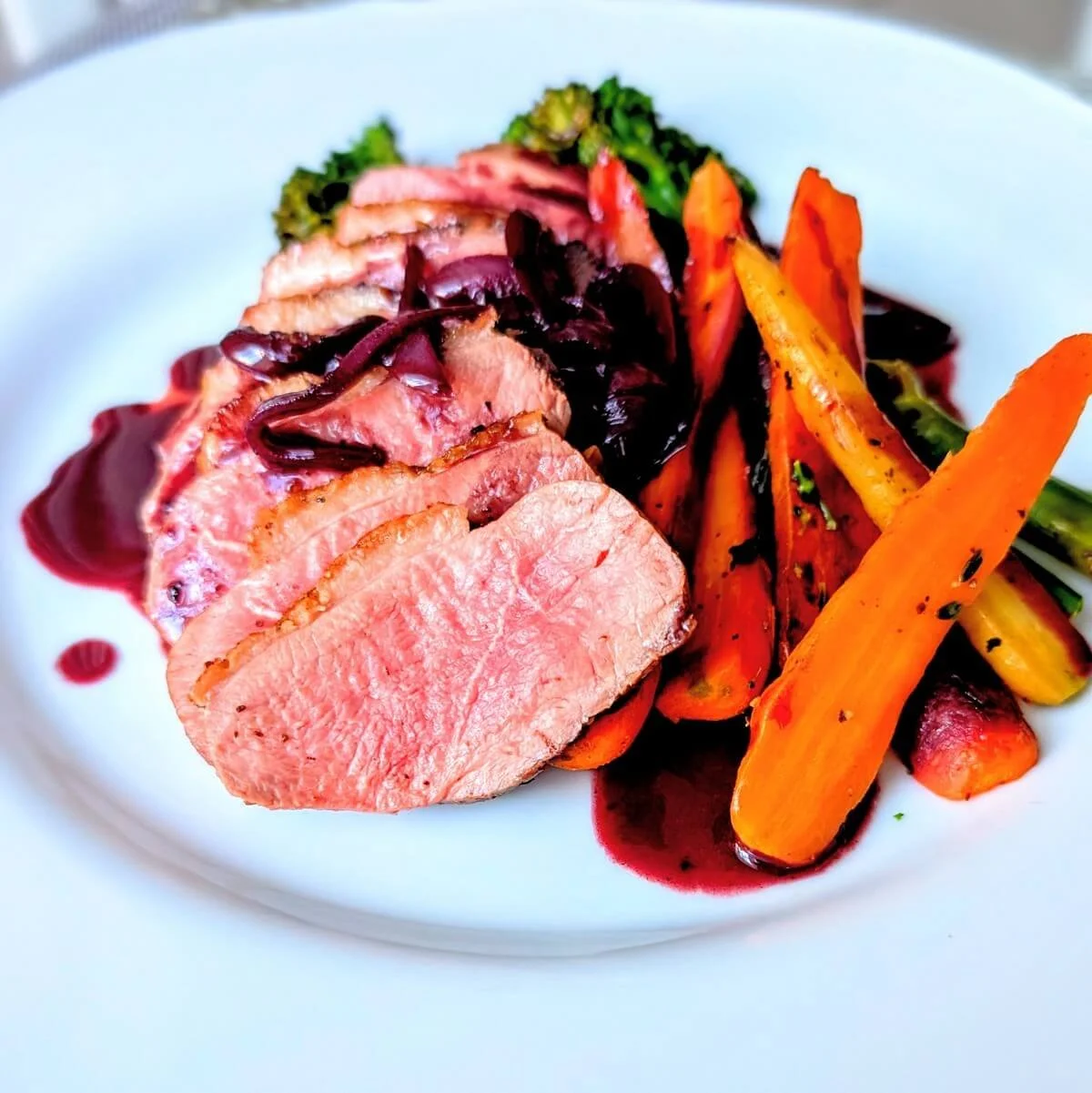 Perfectly cooked medium-rare duck breast slices on a plate with duck pan sauce, carrots, and broccolini