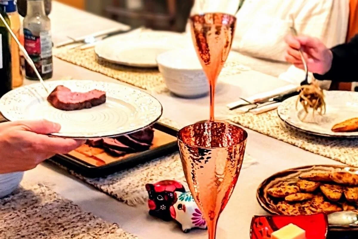 Holiday table setting with venison being plated, surrounded by festive sides and copper wine glasses, capturing a cozy Christmas dinner moment