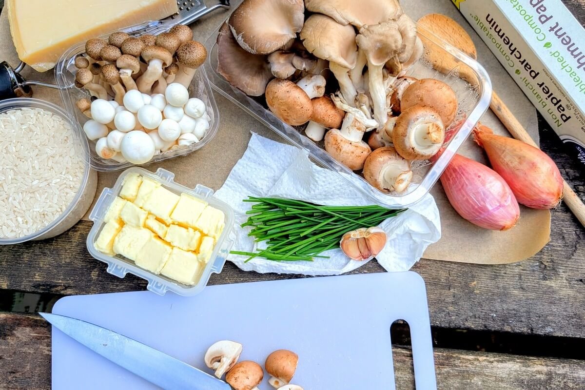 Camping risotto ingredients on a rustic outdoor table with arborio rice, mixed mushrooms, butter, shallots, garlic, chives, and hard cheese