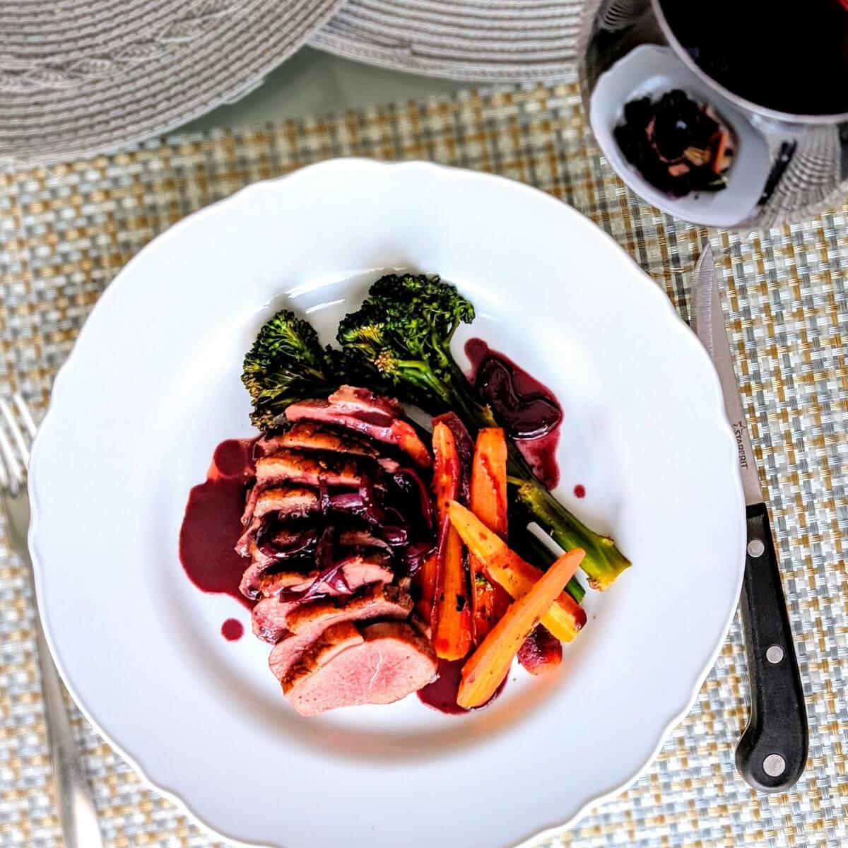 A plate with duck breast, duck pan sauce, and vegetables with a glass of wine