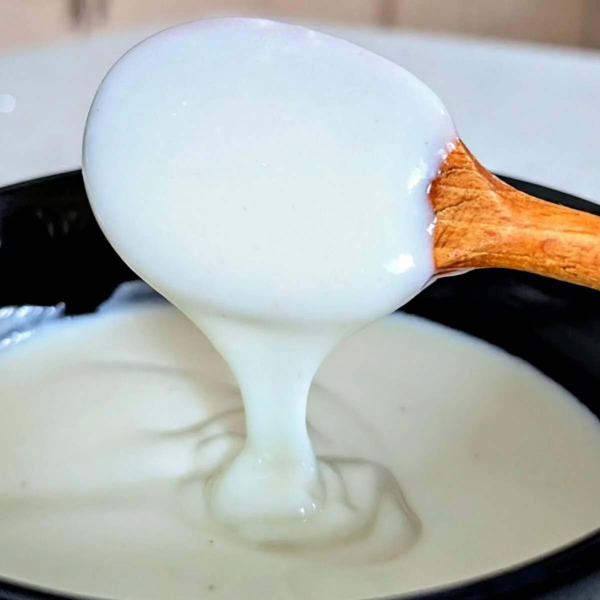 A wooden spoon drizzling bechamel sauce in a bowl with a silky-smooth texture