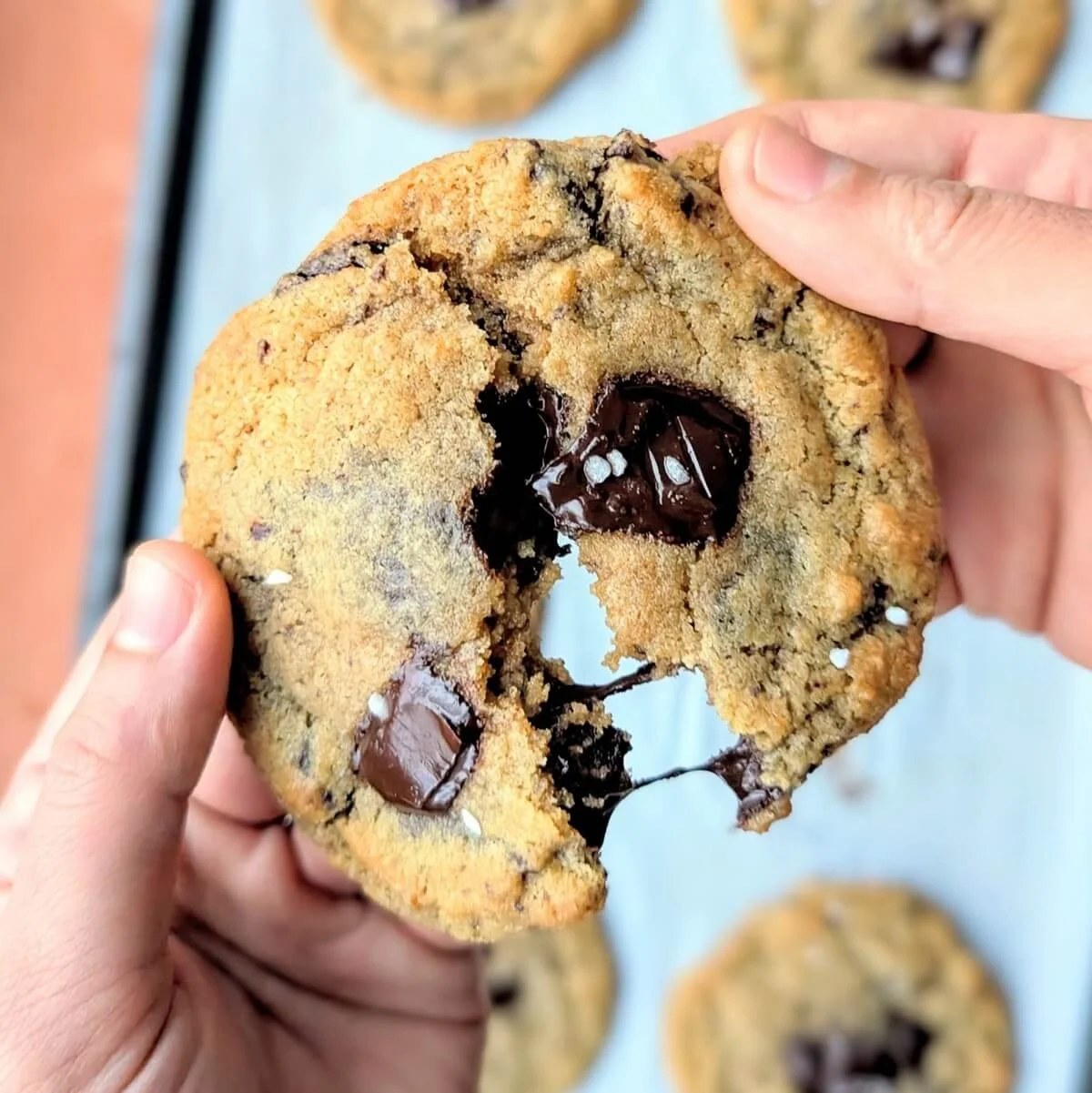 Hands pull apart a gooey dark chocolate chunk cookie for Christmas, revealing melted chocolate and a sprinkle of flaky sea salt, with more cookies visible on a baking tray in the background