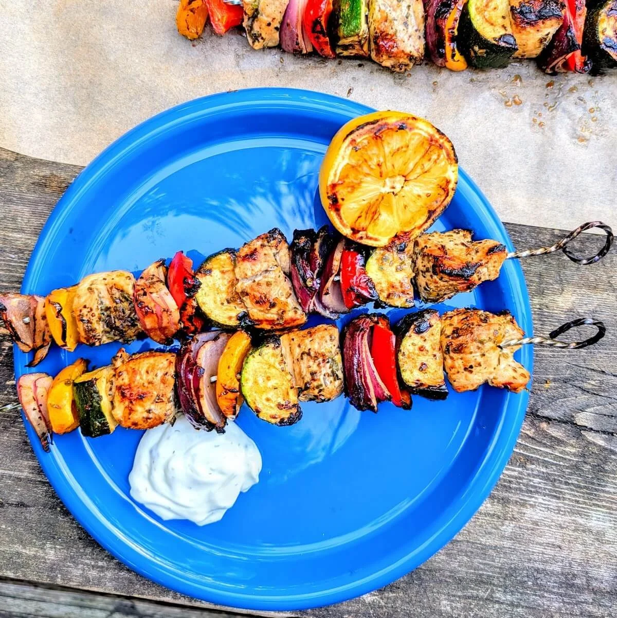 Two Greek chicken skewers served on a blue plate at a campsite with tzatziki and charred lemon on the side