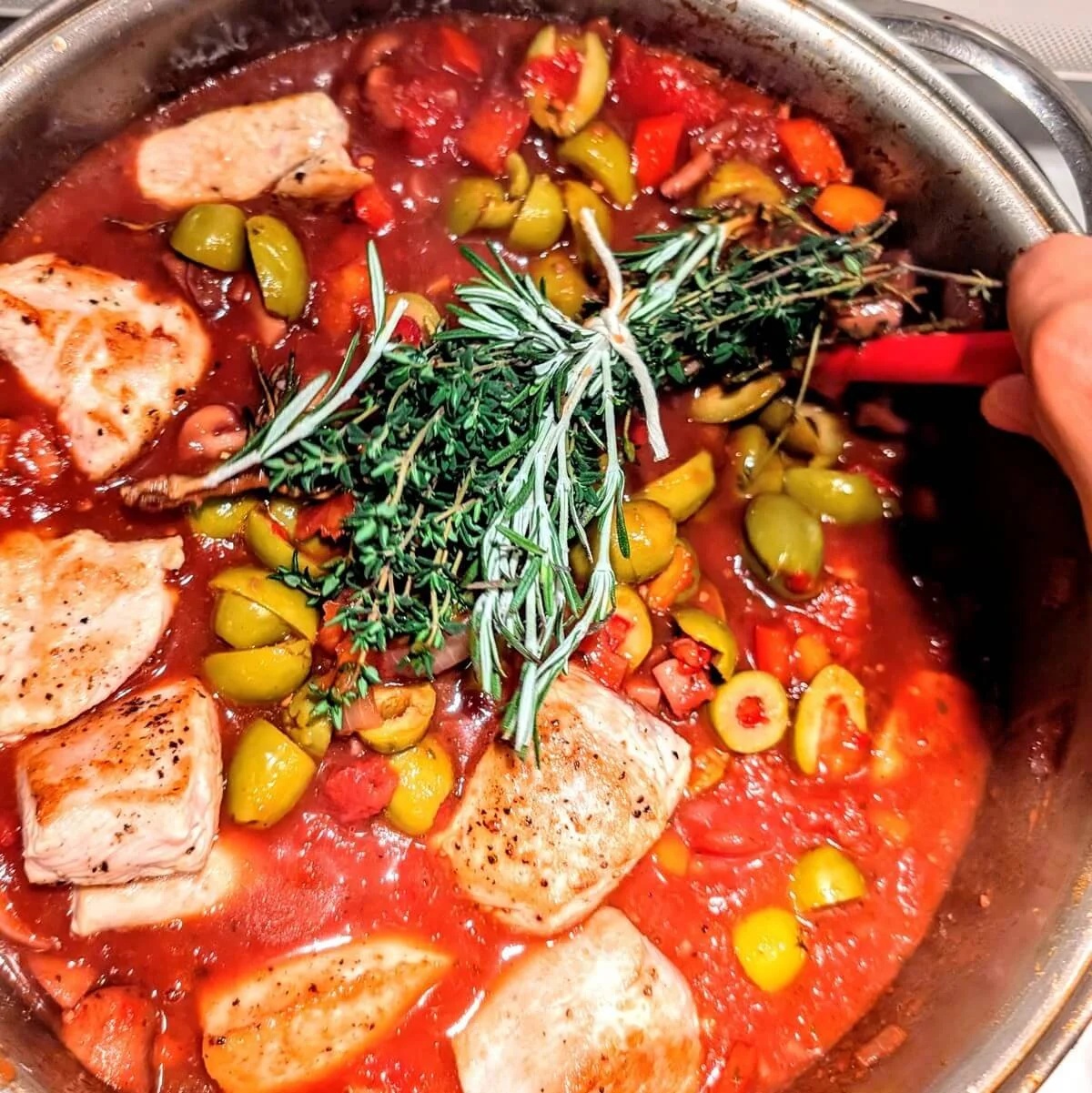 A hand stirring turkey cacciatore as it simmers, with tomatoes, olives, turkey breast, and a visible bundle of rosemary and thyme