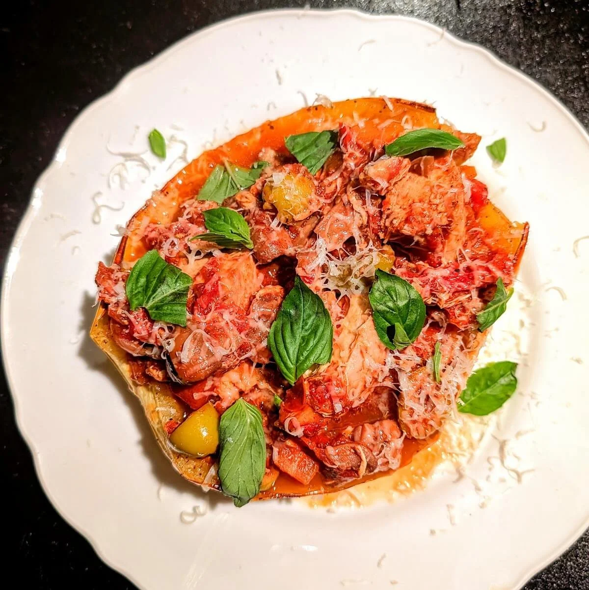 Overhead view of rustic turkey cacciatore recipe served in a roasted spaghetti squash boat, topped with fresh basil, green olives, and grated cheese on a white plate