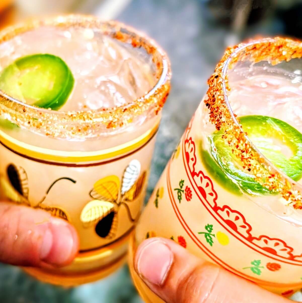 Two festive glasses of mezcalita with chili-lime salt rims, jalapeño slices, and grapefruit-hued cocktails clinked together in a celebratory toast for a Christmas Eve party cocktail