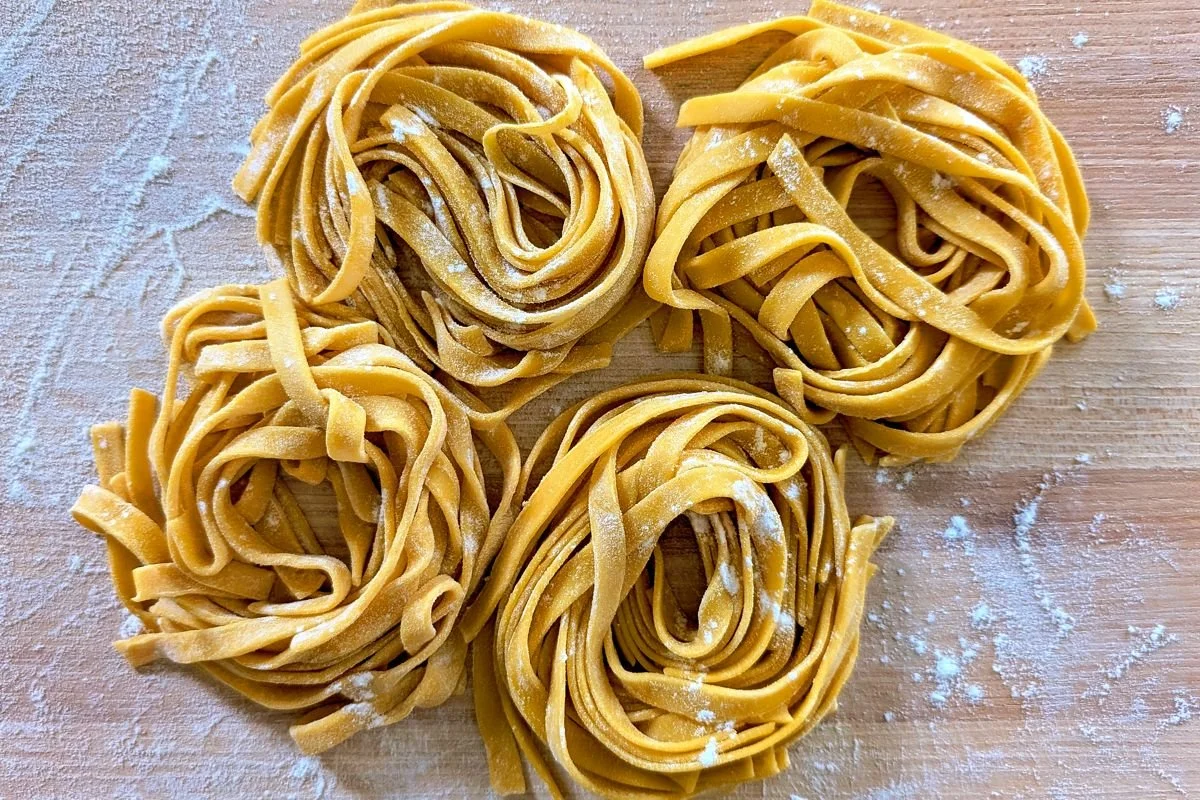 Four nests of fresh egg fettuccine pasta on a wooden cutting board lightly dusted with flour