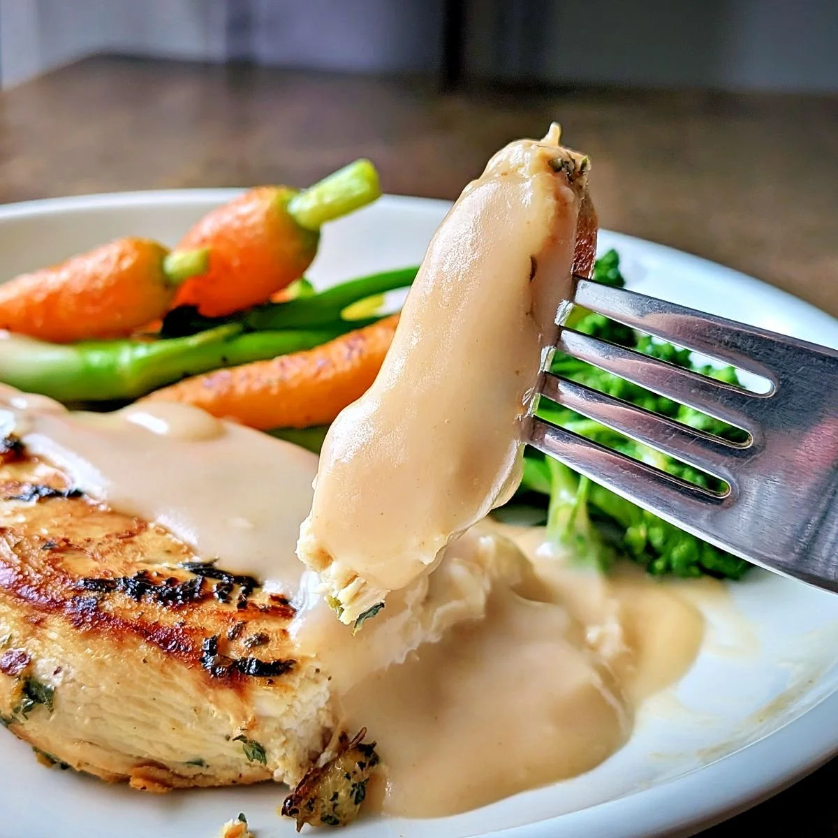 Fork holding a bite of pan-seared chicken coated with creamy velouté sauce over broccolini and carrots