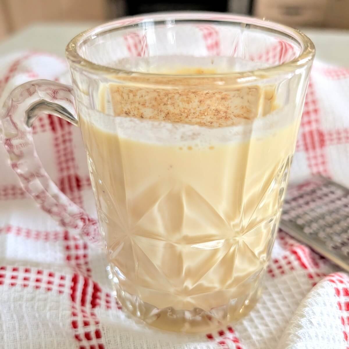 A glass mug of creamy aged eggnog topped with freshly grated nutmeg, set on a festive white and red kitchen towel with a nutmeg grater nearby, ready for Christmas party sipping