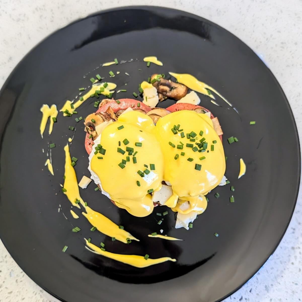 Overhead view of Eggs Benedict on a black plate, topped with homemade immersion blender hollandaise and garnished with chopped chives, with extra sauce artfully drizzled around the plate for Christmas morning brunch