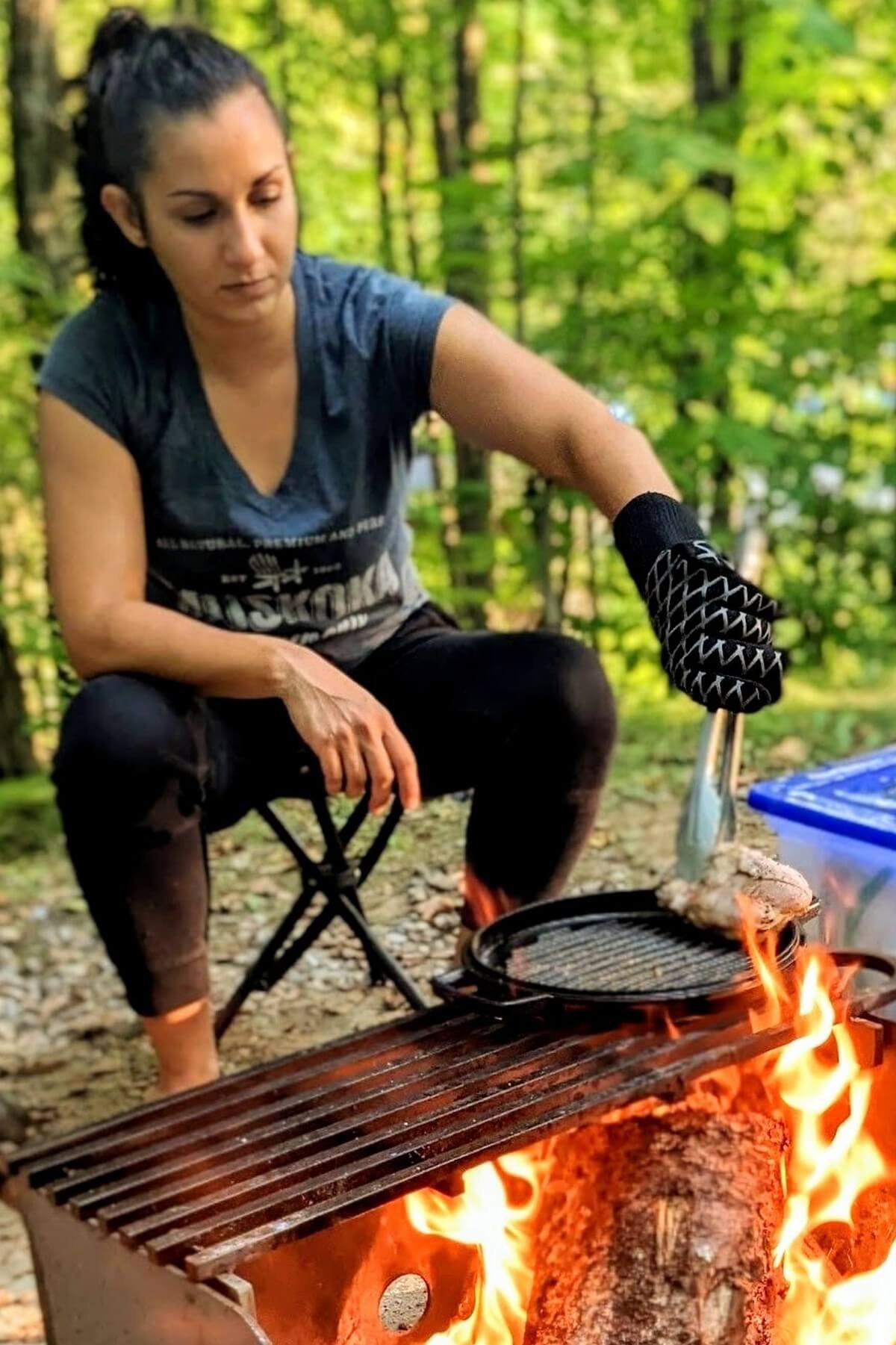 Chef Reem Tiltman cooking over a campfire using a cast iron grill pan, long tongs, and a heat-resistant pit mitt on a fire pit grate