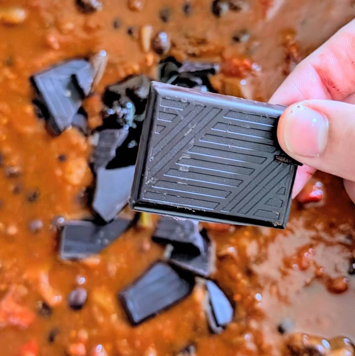Chocolate being added to a pot of chili con carne with braised beef chunks for added depth and complexity