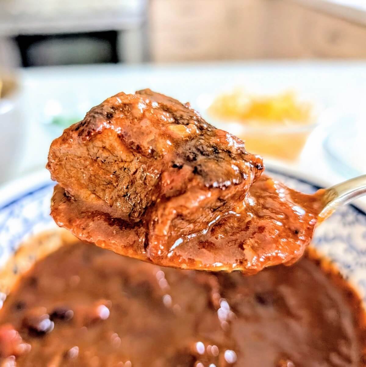 Spoonful of chunky chili con carne with tender braised beef chunk, rich tomato chili sauce, and smoky spices, served in a blue-patterned bowl