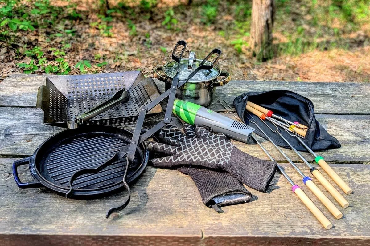 Campfire cooking tools laid out on a wooden picnic table for open fire cooking, including cast iron grill pan, stainless steel pot, firewood tongs, grilling tongs, electric bellows, pit mitts, grill basket, and camp forks