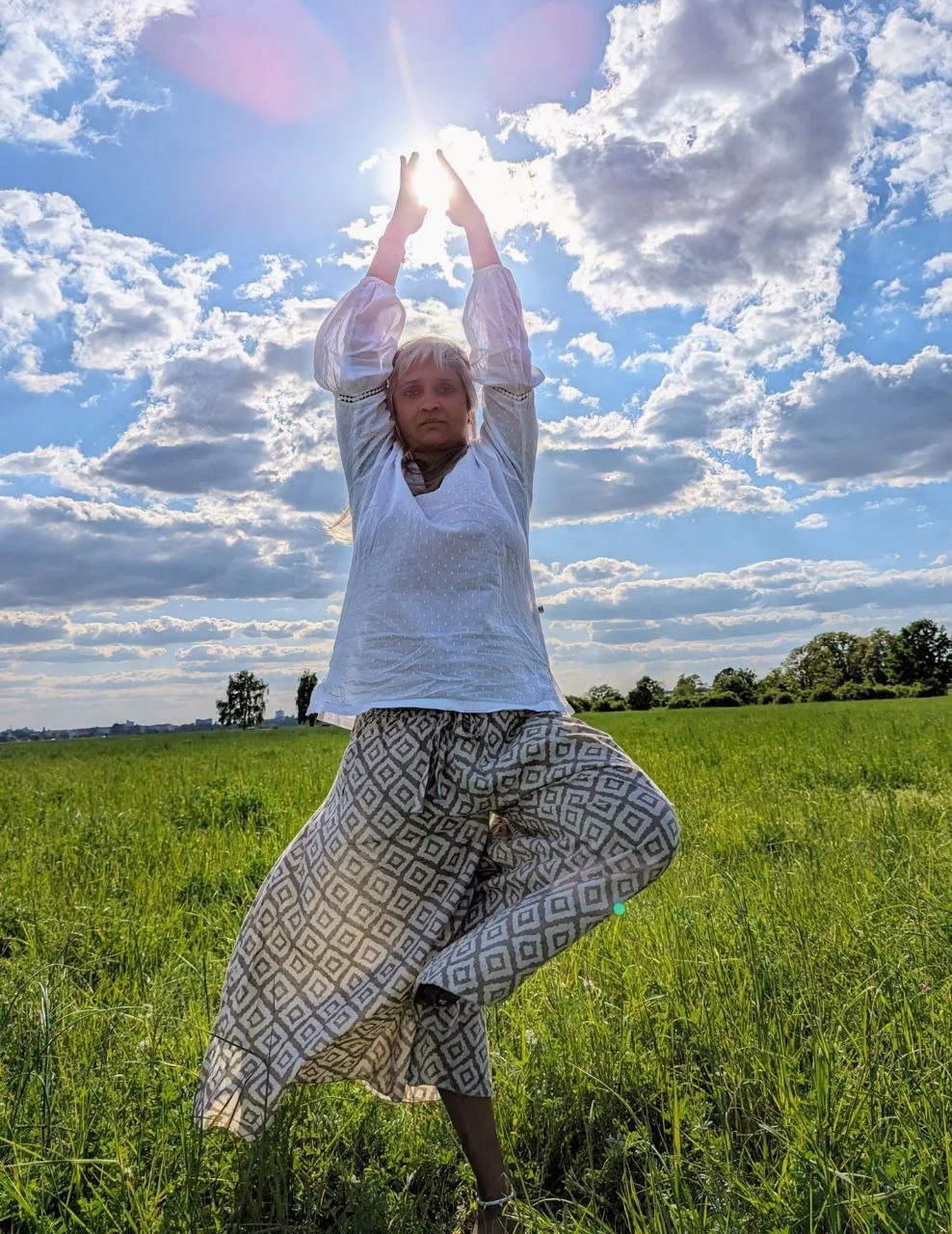 A woman standing in a grassy field with her arms raised toward the sky, touching the sun, against a backdrop of blue sky and clouds.