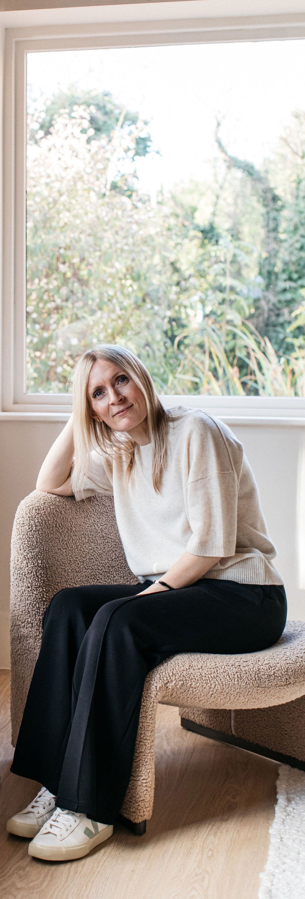 A woman with blonde hair sitting on a plush beige armchair near a large window with a garden view, wearing a cream sweater, black pants, and white sneakers, smiling softly.