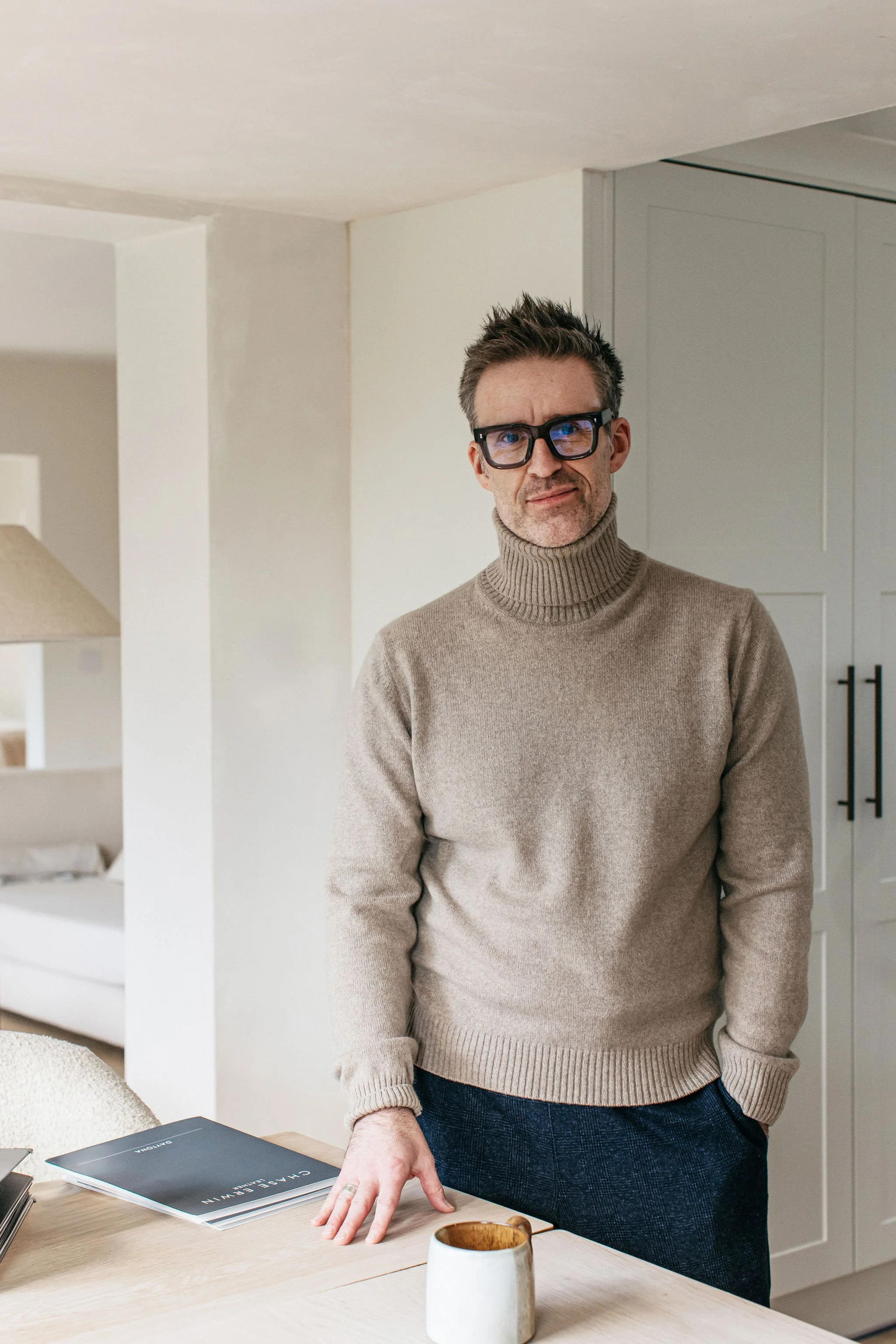 A man with dark hair, glasses, and a beard, wearing a beige turtleneck sweater, standing in a modern, minimalistic room with white walls and a light-colored wooden table.
