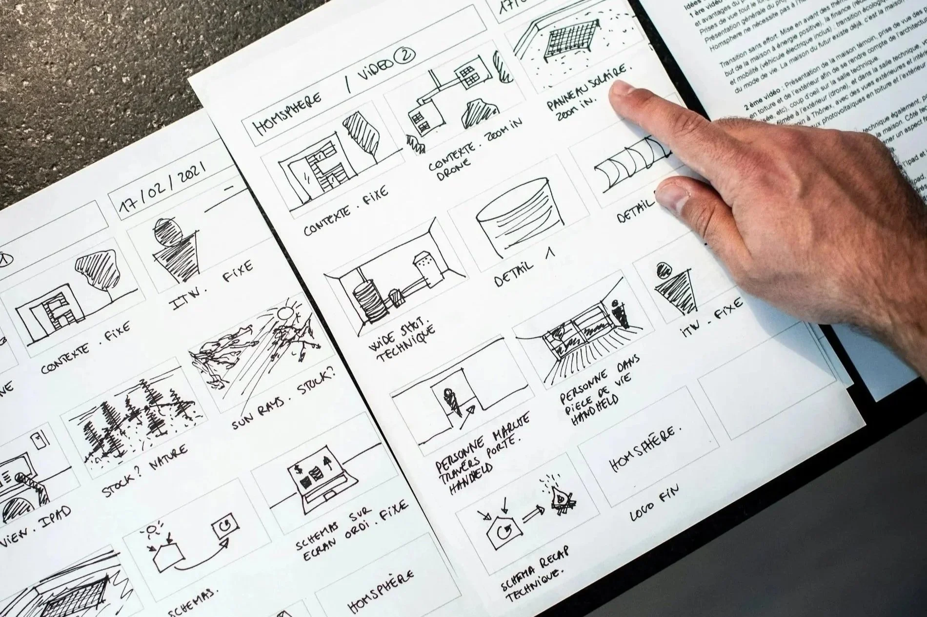 Hand pointing at a page with sketches and notes about a project, including diagrams of buildings, technical details, and environmental themes, on a printed sheet.