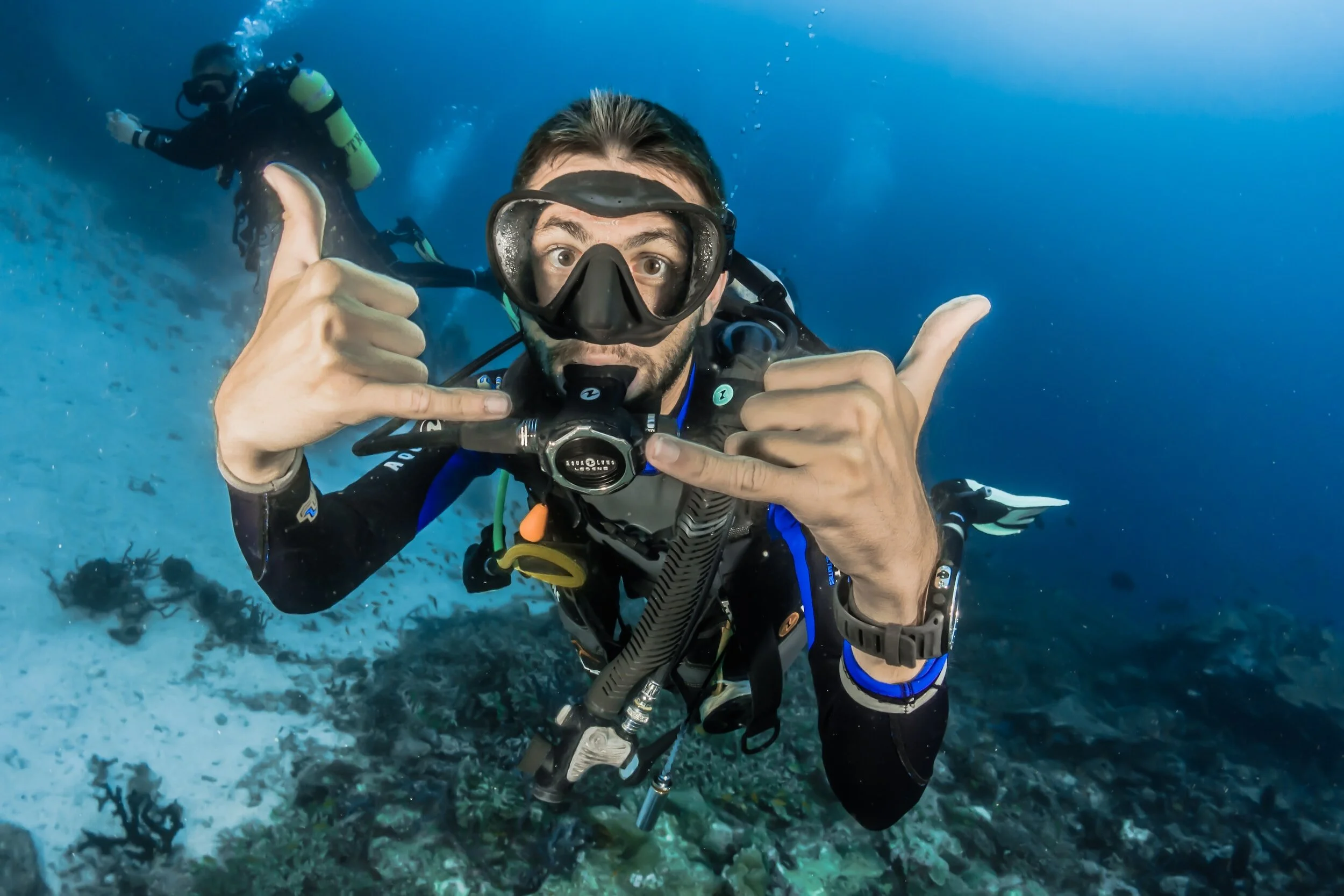 A scuba diver underwater showing a thumbs-up gesture and making a shaka sign, with another diver in the background.
