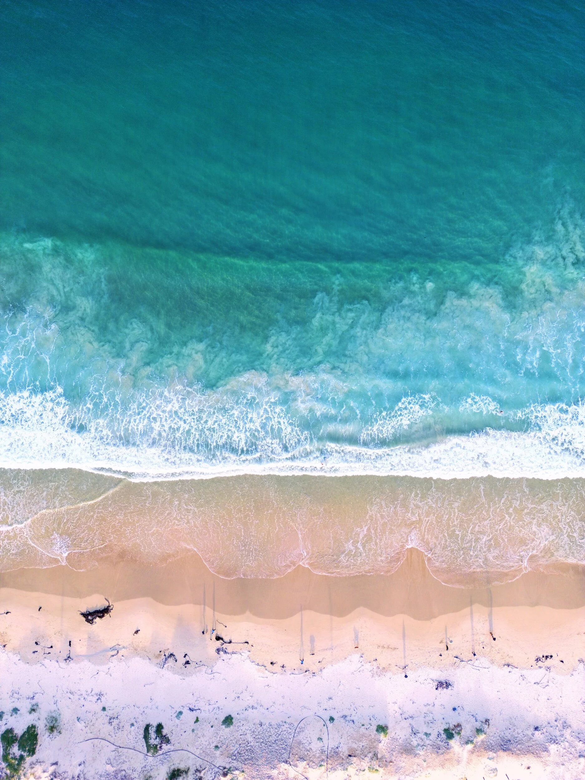 Aerial view of a beach showing turquoise ocean waves, sandy shoreline, and some debris and vegetation along the edge.