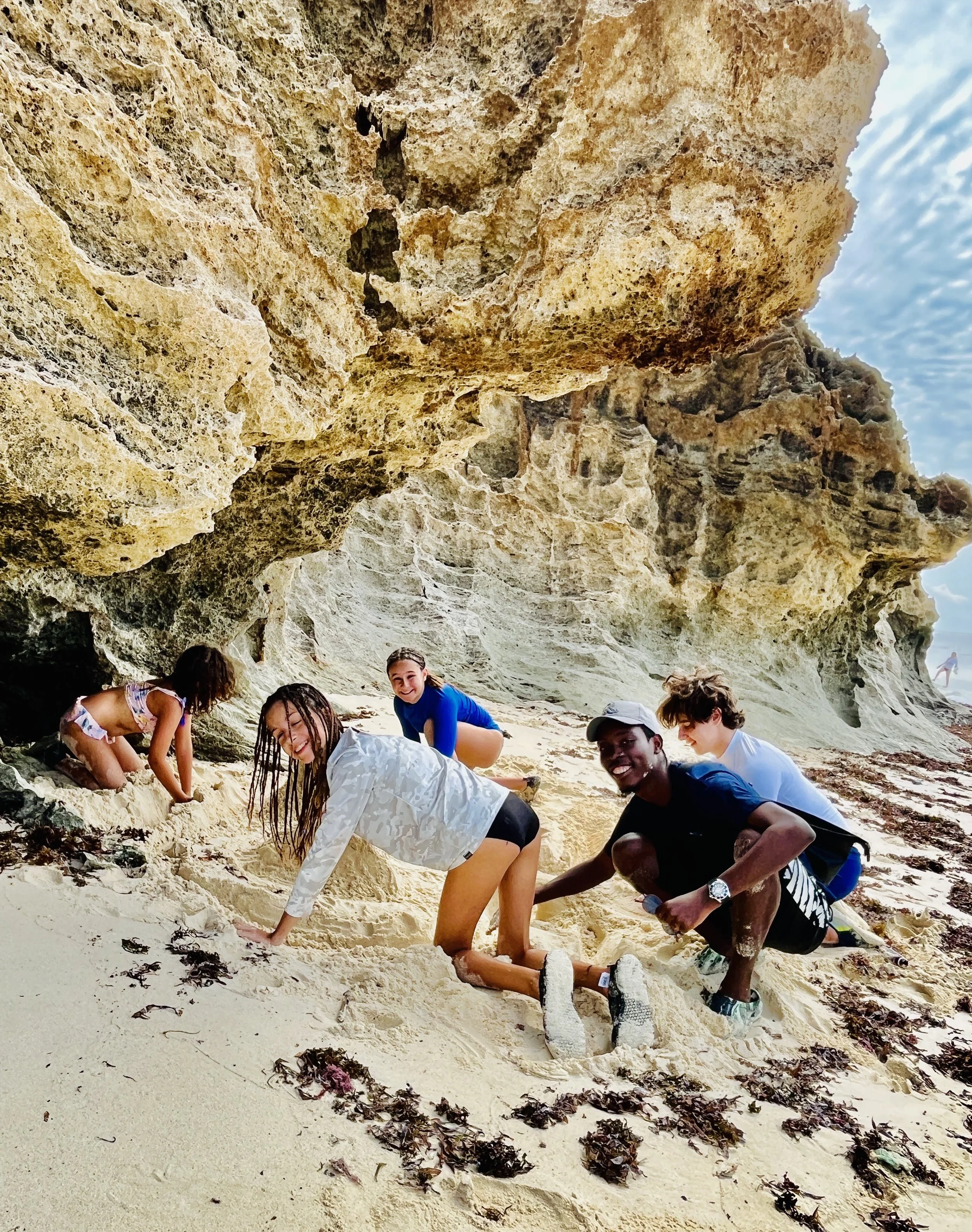 A group of five people, including children and adults, playing and exploring on the sandy beach near a rocky cliff with some seaweed scattered around.