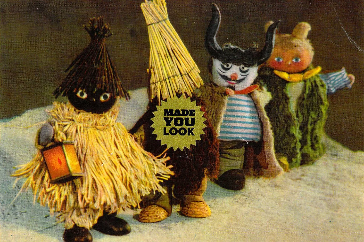 Three small puppets dressed in colorful costumes standing together, with a dark background. One puppet holds a small lantern, another has a sign that says "Made You Look," and the third has curly hair and a scarf.