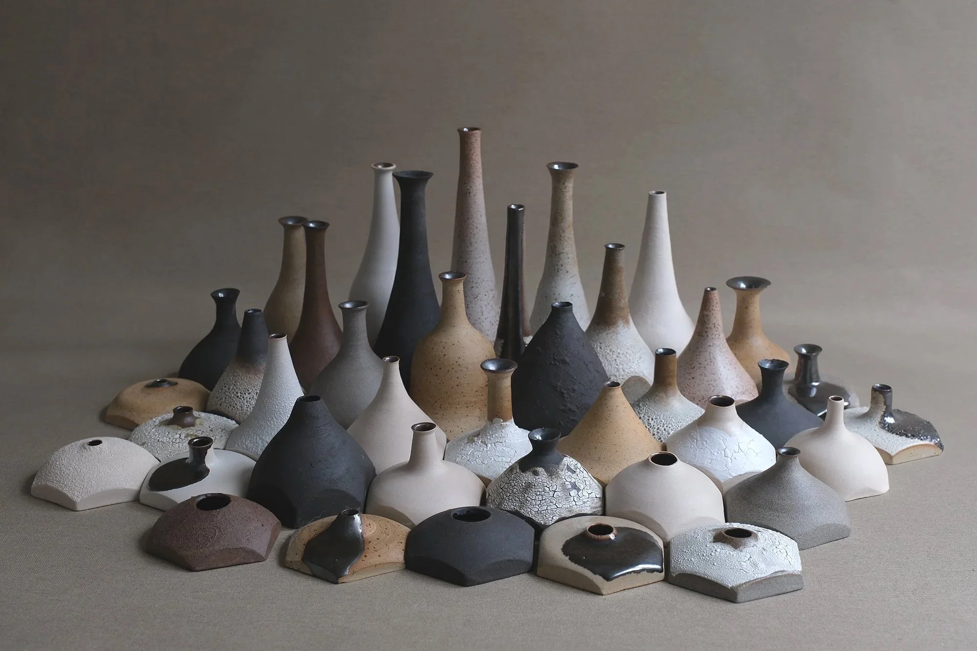 Collection of various ceramic vases and pottery in neutral tones arranged on a flat surface with a plain background.