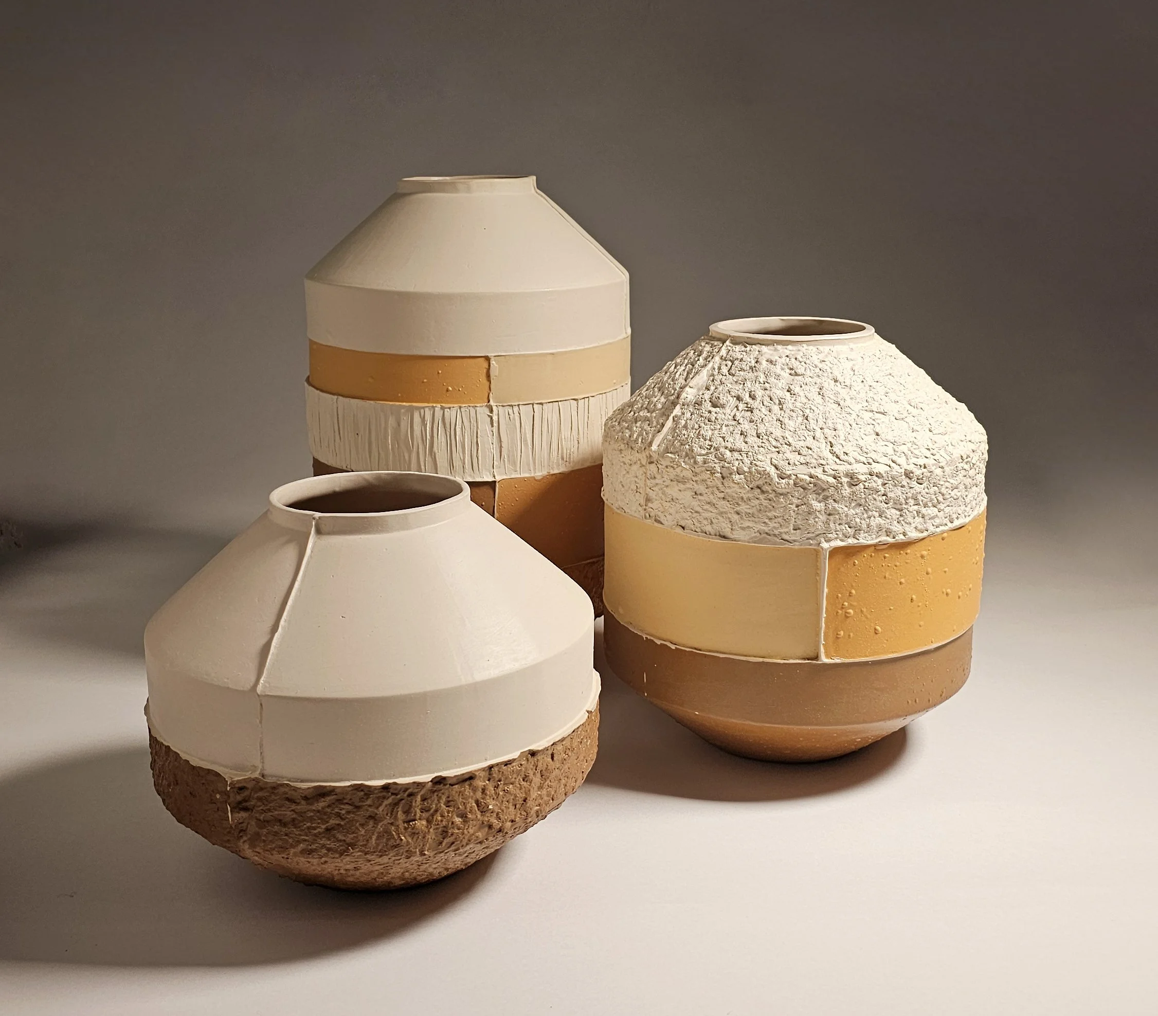 Three ceramic vases with unique textures and color bands, arranged on a white surface against a gray background.