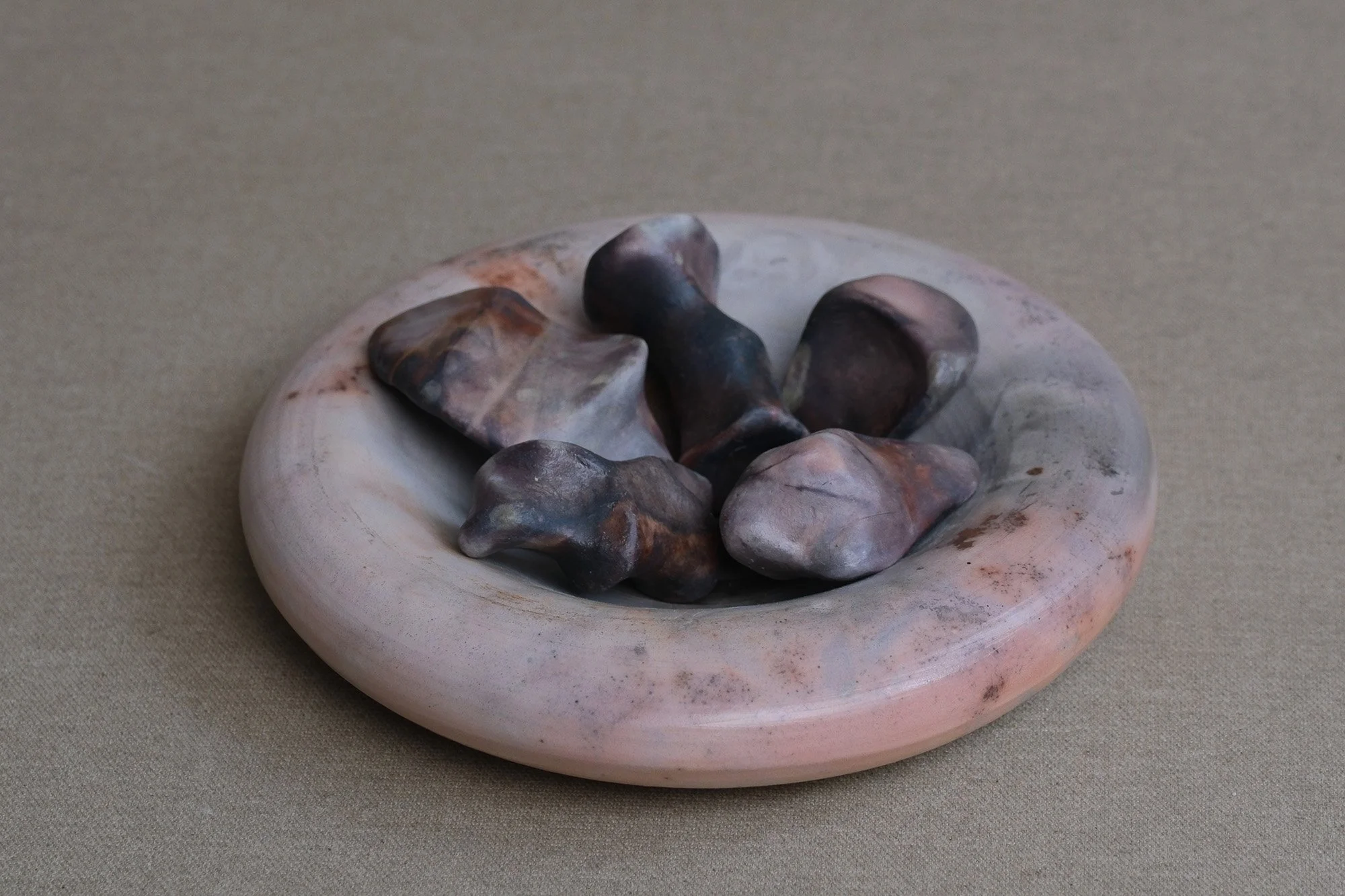 A round, shallow bowl with a swirled pink and white pattern, containing several dark, irregularly shaped stones, set against a beige fabric background.