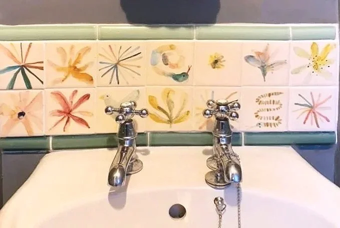 Bathroom sink with colorful flower and bird tiles behind two chrome faucets.