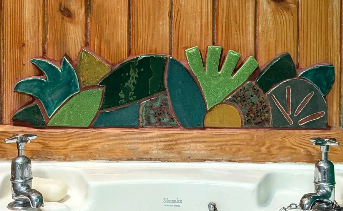 Colorful mosaic tile art of various green plants and leaves on a wooden wall above a white sink.