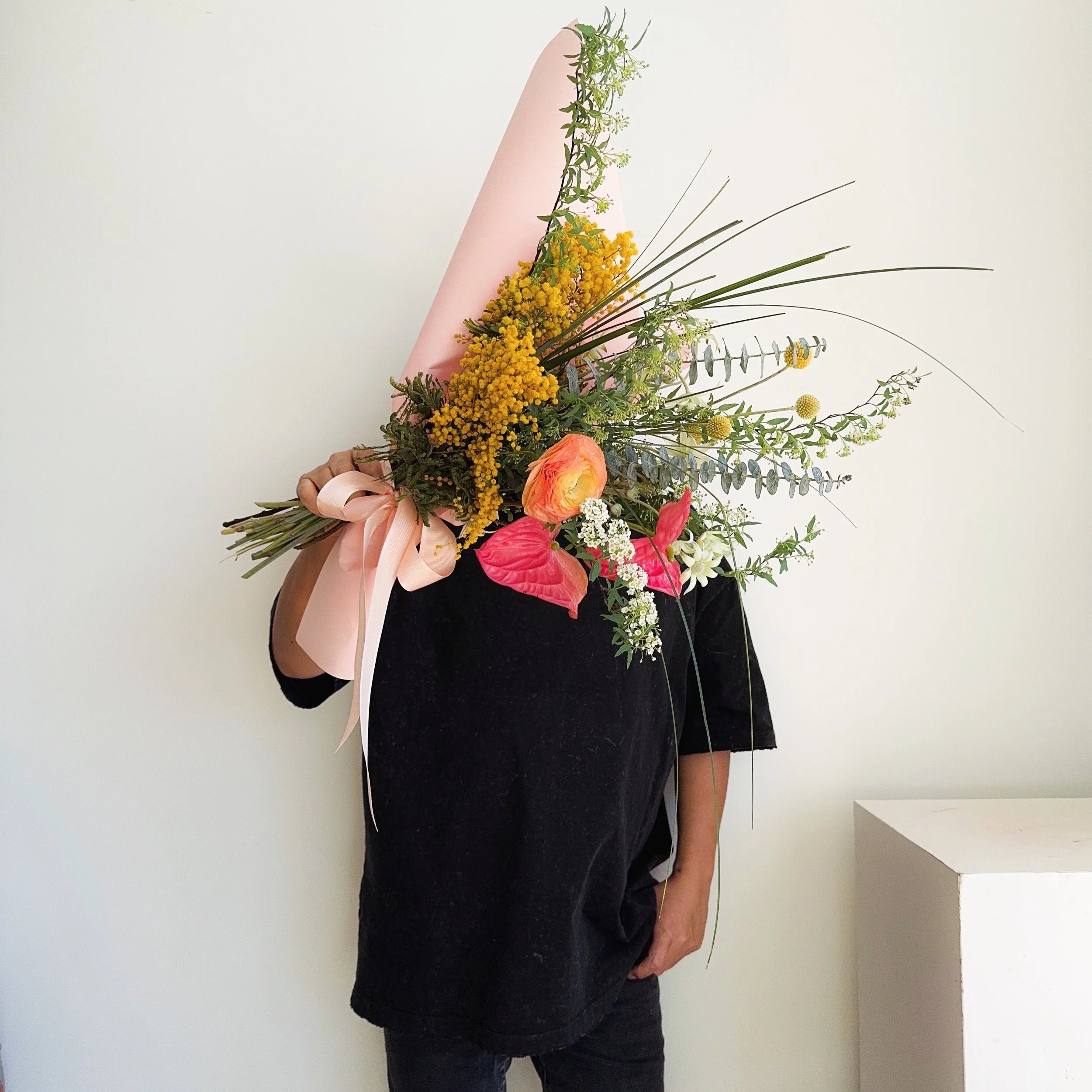 Person holding a large bouquet of colorful flowers, covering their face, against a plain white wall.
